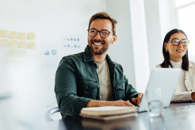 A smiling employee in a green shirt and glasses sits in a meeting with a colleague